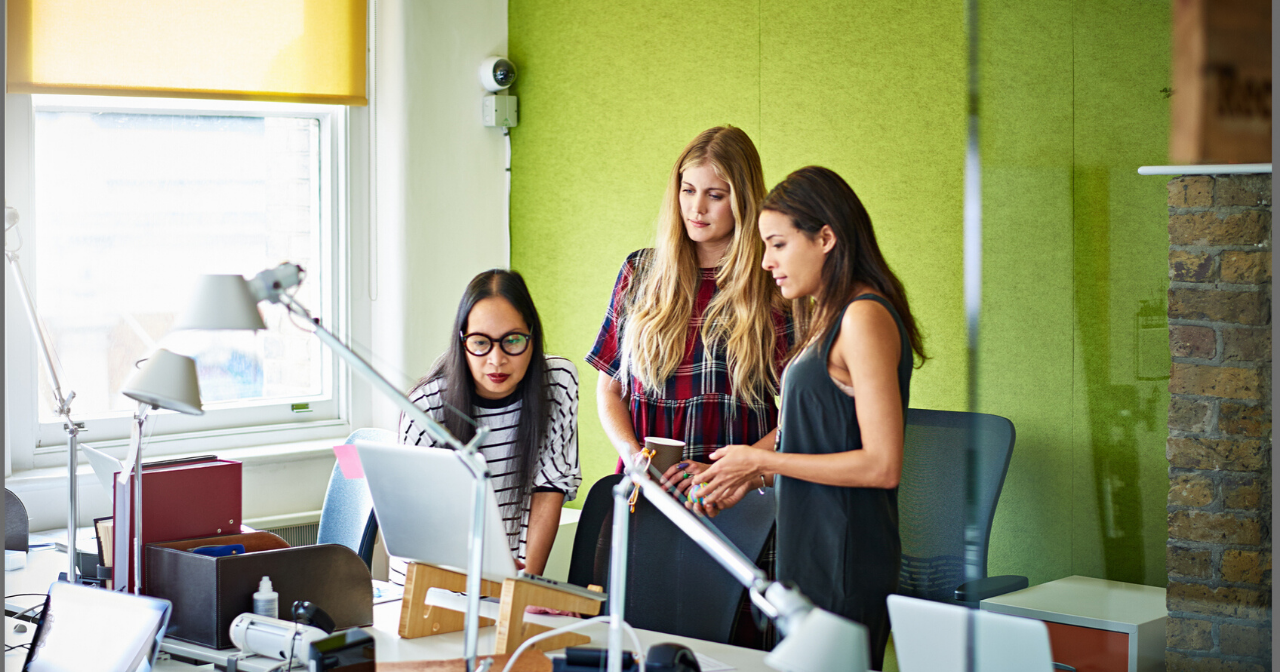 Three women gather around a computer in an office