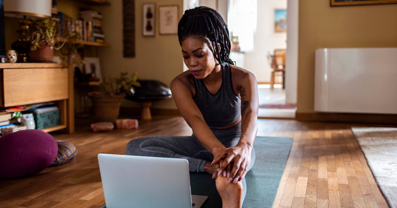 A woman stretches on a yoga mat while looking at her computer. 