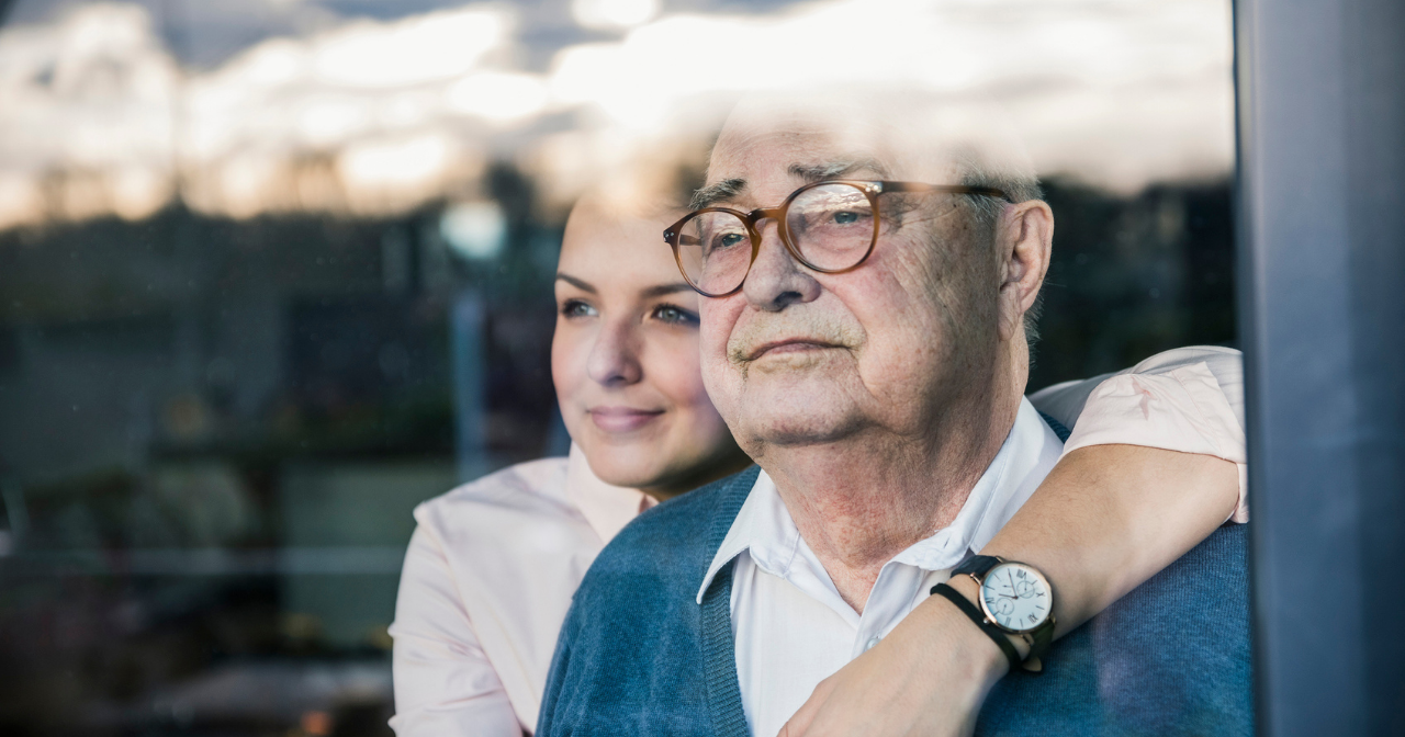 A woman hugs her grandfather from behind as they both look out a window. 