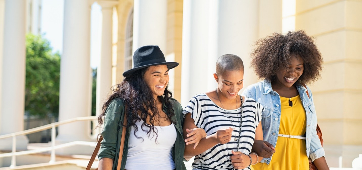 Three young woman link arms and laugh. 