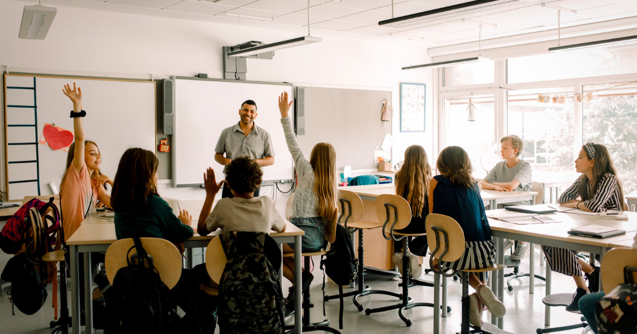 An engaged middle school classroom with a teacher at the head and several hands raised. 