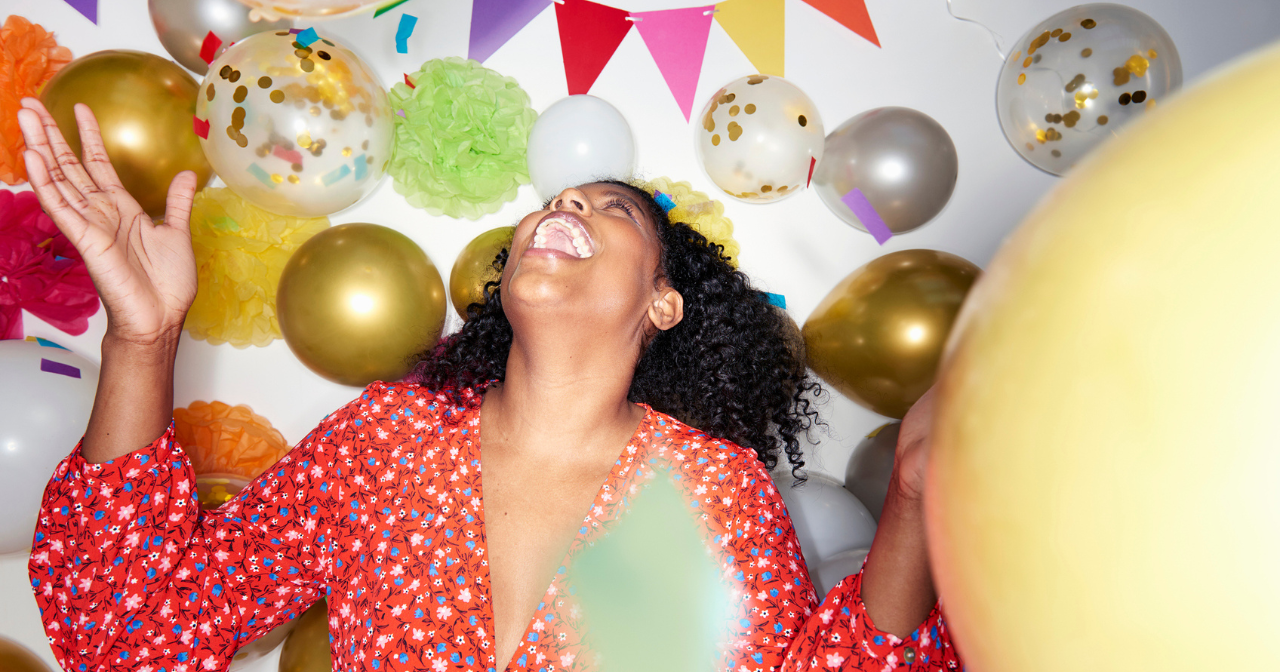 A woman stands and throws her head back laughing while surrounded by balloons. 