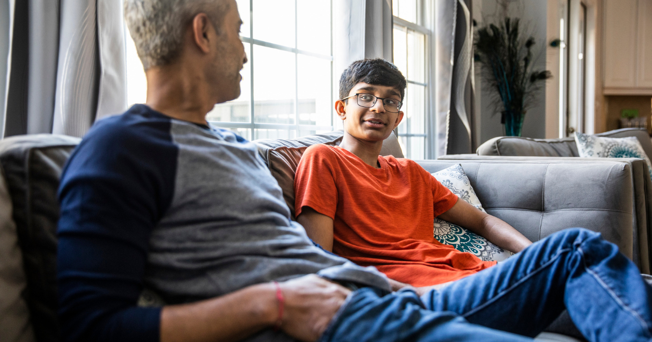 A father and his young teen son sit on the sofa and talk. 