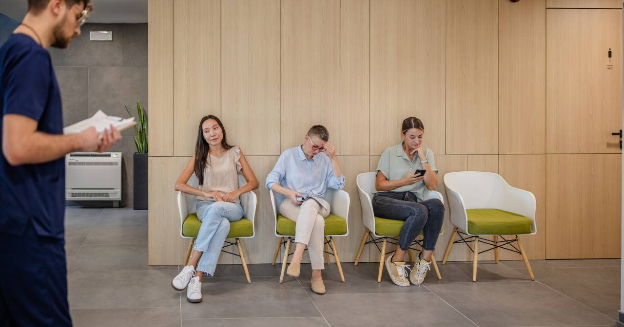 A clinic waiting room with three women waiting in chairs as a health care provider walks past. 