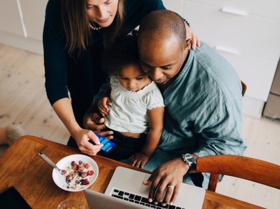 A father sits with his child on his lap at the breakfast table while the child's mother leans over his shoulder to look at his laptop screen. 