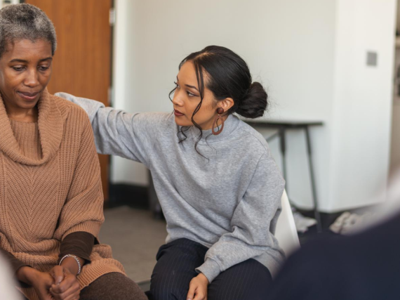 One Black woman puts an arm around another during group therapy.