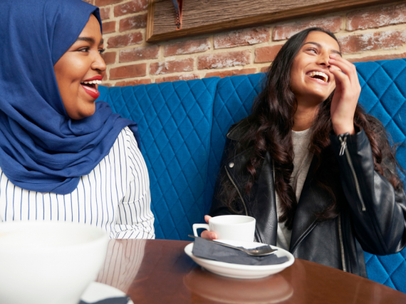 Two women sit at a table in a cafe and laugh over cups of coffee. 