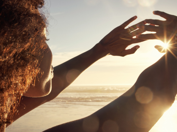 A Black woman stands on the beach and holds her hands up so she can look at the sunset. 