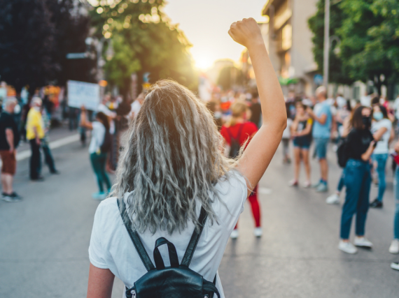 A photo of the back of a young woman at a protest with her fist raised. 