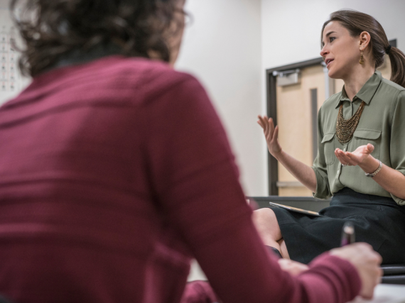 A teacher sits at the front of the classroom and talks to her students as they take notes. 