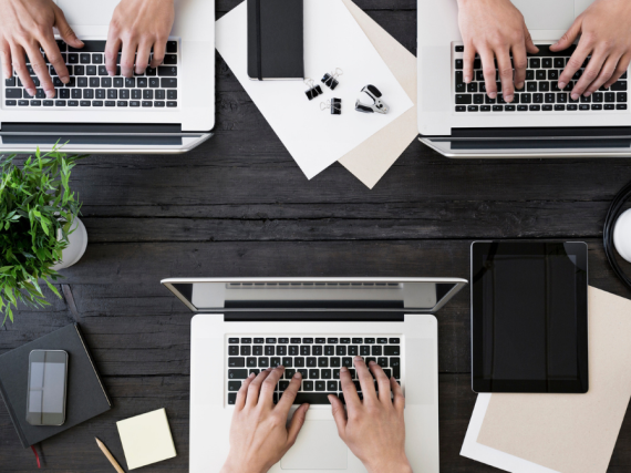 Three sets of hands on three laptops on a large table. 