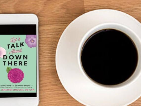 An image of a phone and a cup of coffee on a table. The cover of the book 'Let's Talk About Down There' is on the phone. 