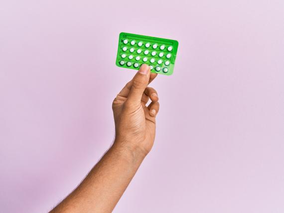 A hand holds a generic pill pack against a purple background. 