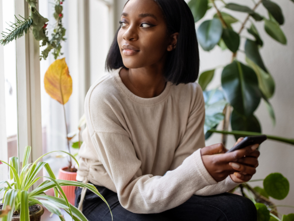 A Black woman sits next to a window and looks out contemplatively. 