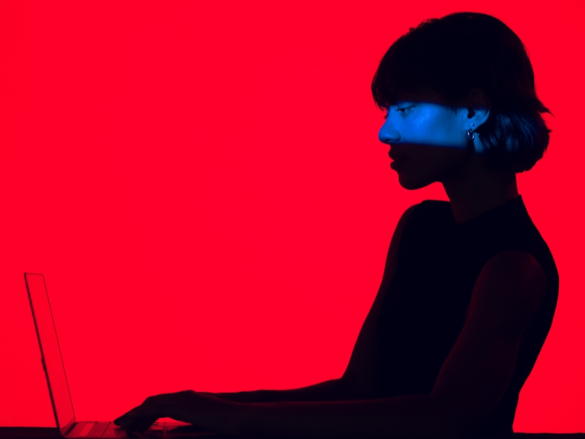 A woman in shadow types on a laptop. The background is bright red.